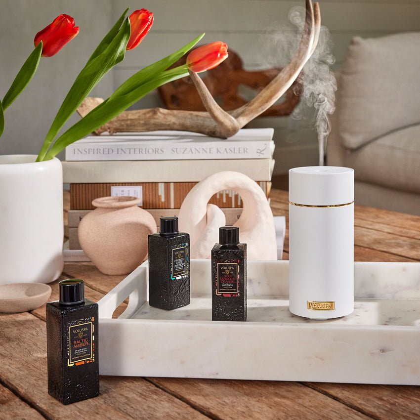 A Voluspa White Ultrasonic Fragrance Oil Diffuser emits mist on a marble tray beside three black fragrance bottles with gold accents. Red tulips in a vase, books, and decorative items adorn the background on a wooden table.