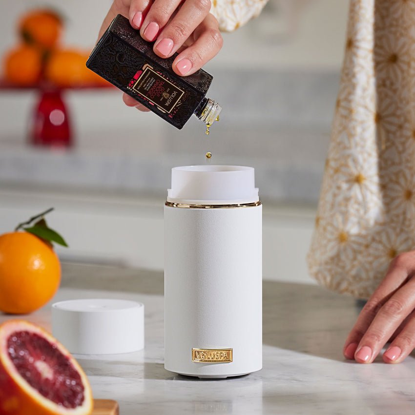 A person fills a Voluspa White Ultrasonic Fragrance Oil Diffuser Device on a marble counter. Blurred fresh oranges and a cut blood orange add color, while a decorative pattern adorns the persons sleeve.