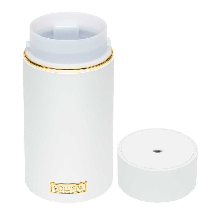 The Voluspa White Ultrasonic Fragrance Oil Diffuser, a cylindrical vessel with a gold accent and removable lid, rests on a white background. The label reads Voluspa and the lid is placed beside it.