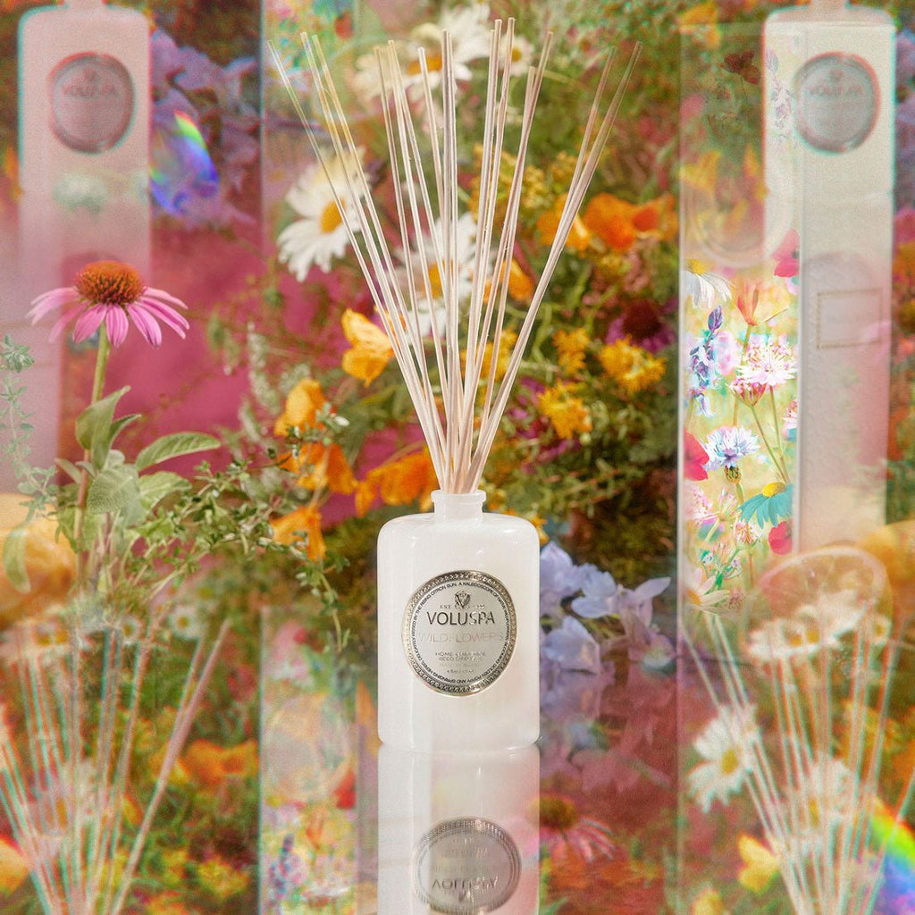 The Voluspa - Wildflowers 172ml Reed Diffuser, featuring thin sticks in a white bottle, is surrounded by daisies and golden poppies. The vibrant floral backdrop creates a kaleidoscope effect, enhancing the fragrance experience.