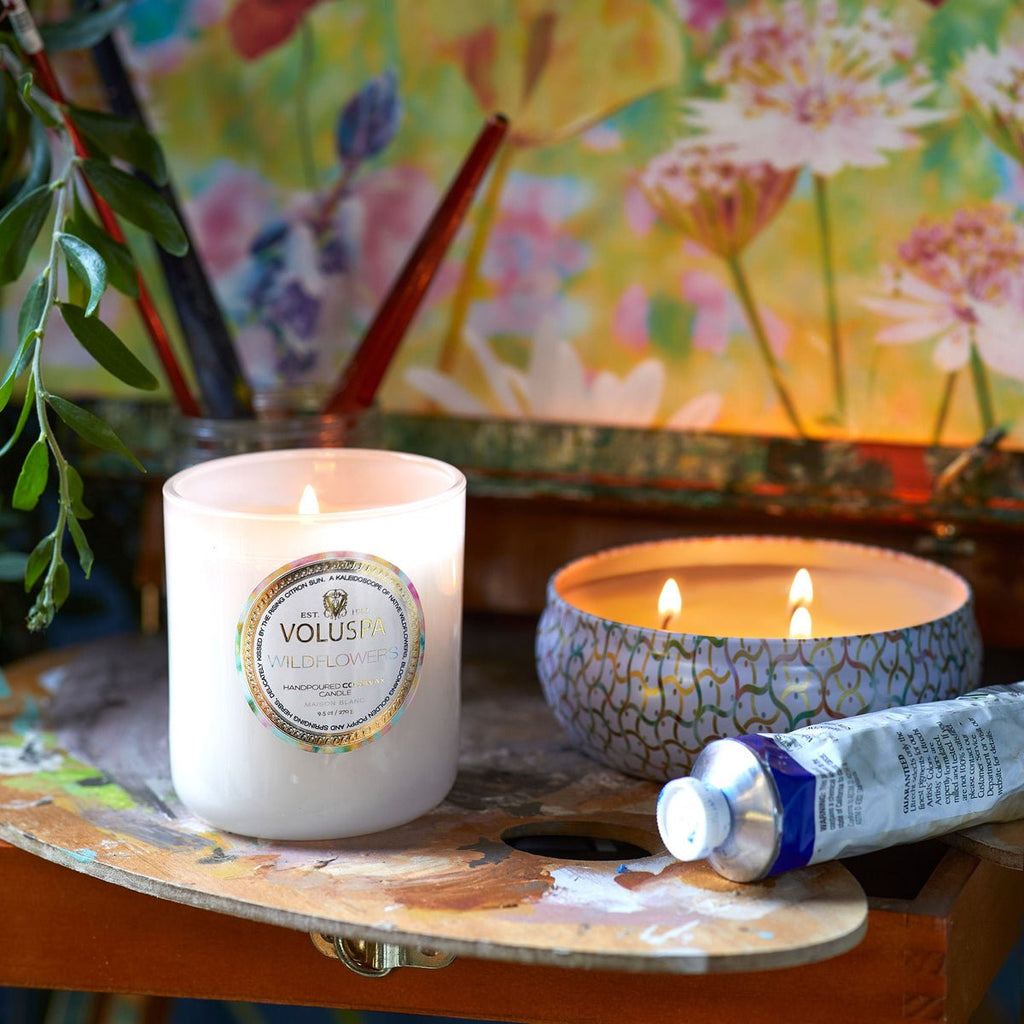 A lit Voluspa Wildflowers Classic 9.5oz candle sits on an artists palette beside a small blue patterned coconut wax bowl candle. Nearby are paintbrushes, a paint tube, and a vibrant floral painting in the background.