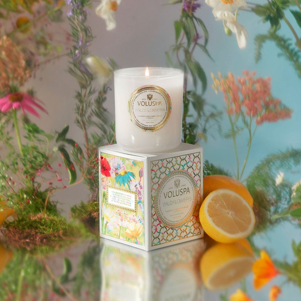 A lit Voluspa - Wildflowers Classic 9.5oz Candle, crafted from a clean-burning coconut wax blend, rests on a decorative box with wildflowers and lemon halves on a reflective surface, creating a bright and fresh floral ambiance.