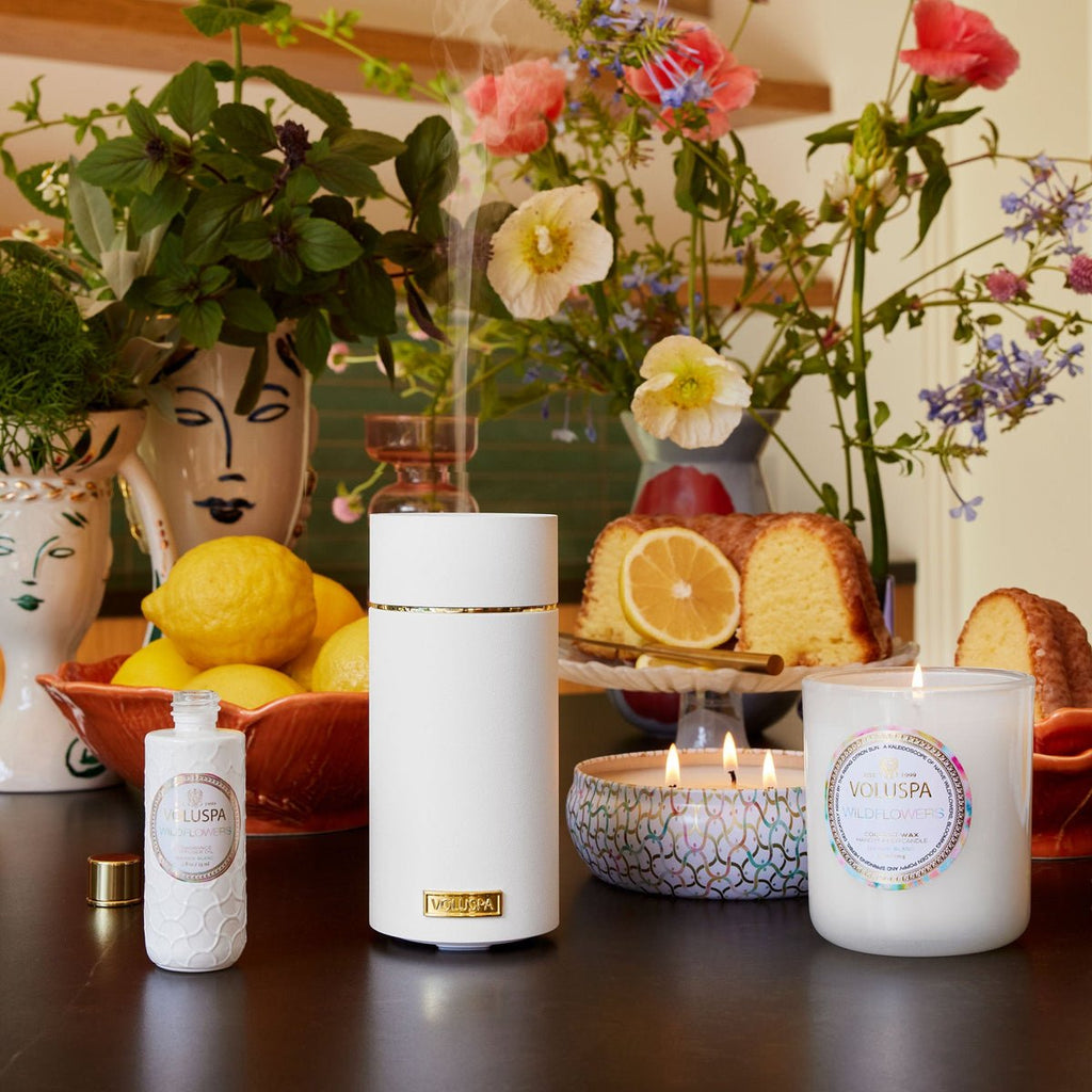 A cozy table setting features a Voluspa Wildflowers Fragrance Diffuser, a Voluspa Fragrance candle, lemon pound cake with lemon slice, and lotion bottle. Colorful flowers in artistic vases and a bowl of lemons enhance the vibrant backdrop with the allure of golden poppies.