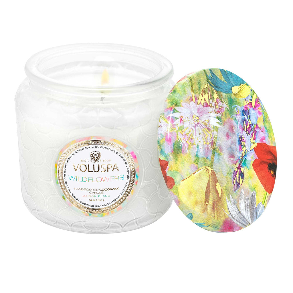 The Voluspa - Wildflowers Petite Jar Candle 4.5oz features a coconut wax blend in a textured glass jar, releasing a delightful floral scent. Its colorful floral-patterned lid complements the warm glow of the lit candle.