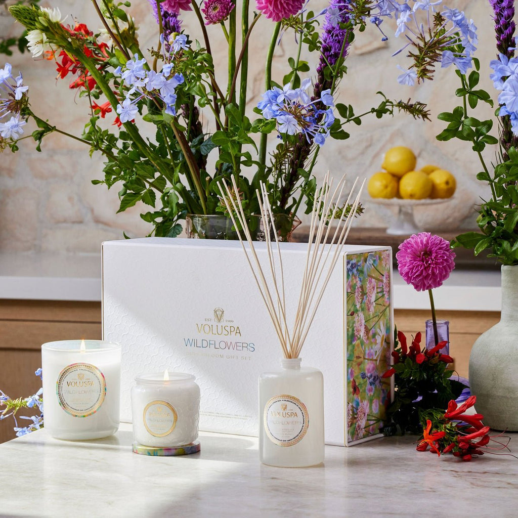 Floral-scented candles and a reed diffuser rest on a marble surface. Behind them, a large white Voluspa - Wildflowers Superbloom Gift Set Candles + Diffuser box sits, adorned by native wildflowers, golden poppies, and citrus lemons.