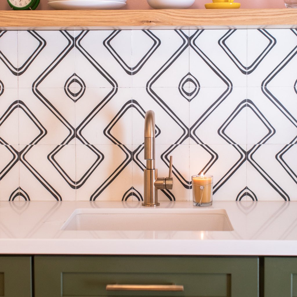 A brass faucet kitchen sink stands against geometric black-and-white tile backsplash. A Votivo - Pink Mimosa Aromatic Candle flickers on the countertop, while a wooden shelf above holds white dishes and a small green clock. The cabinetry is painted green.