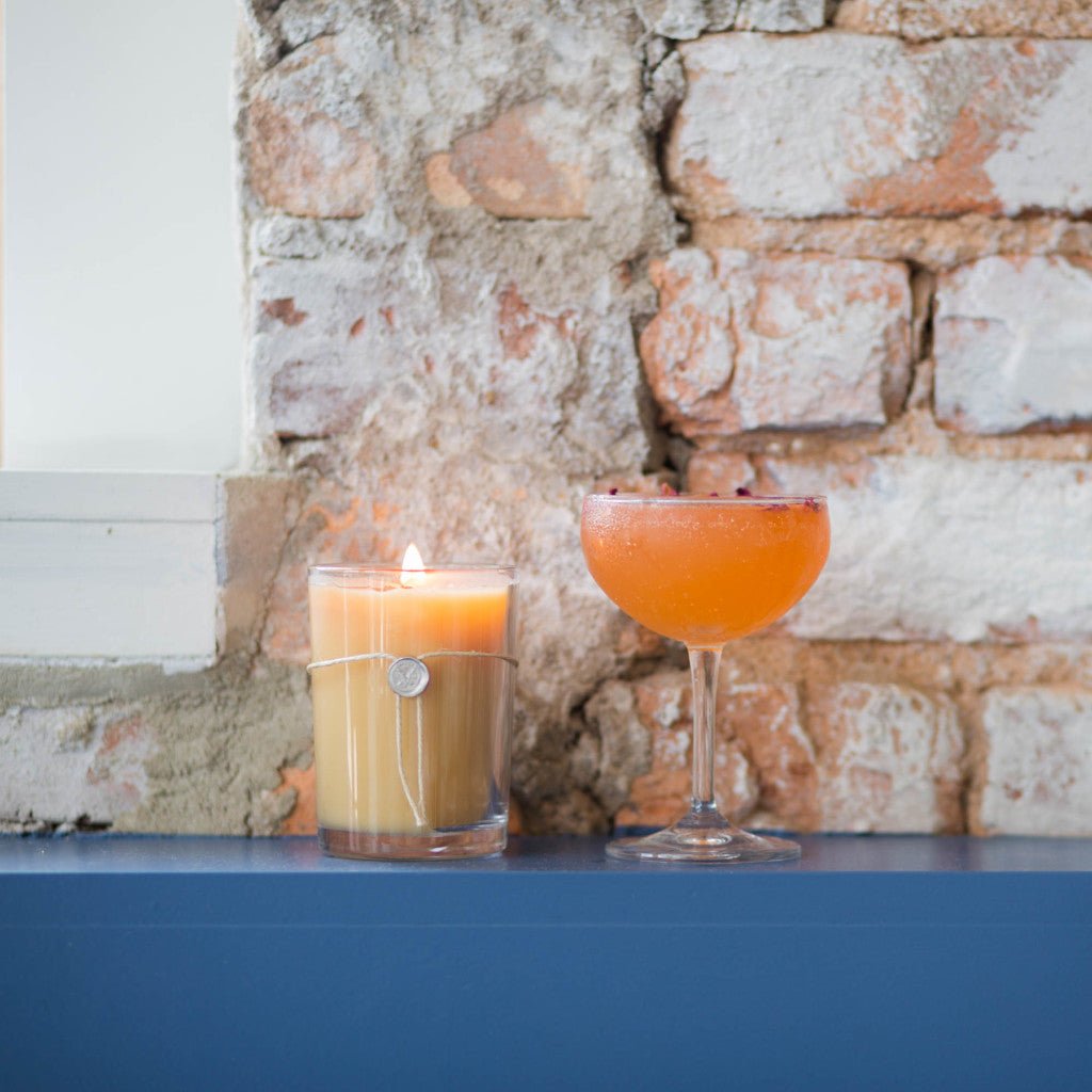 A Votivo - Pink Mimosa Aromatic Candle and an orange cocktail in a coupe glass rest on a blue surface against a rustic brick wall, filling the air with the subtle scent of Votivos pink mimosa fragrance.