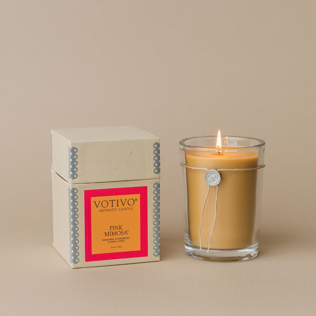 A lit Votivo - Pink Mimosa Aromatic Candle in a clear glass holder sits beside a decorative box against a neutral beige background, crafted with soy-blend wax. The elegance of both items is enhanced by the Pink Mimosa fragrance.