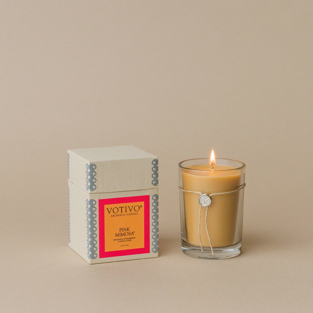 A Votivo - Pink Mimosa Aromatic Candle, crafted from soy-blend wax with a silver charm, sits in a clear glass holder beside its beige box, radiating elegance against a neutral backdrop.