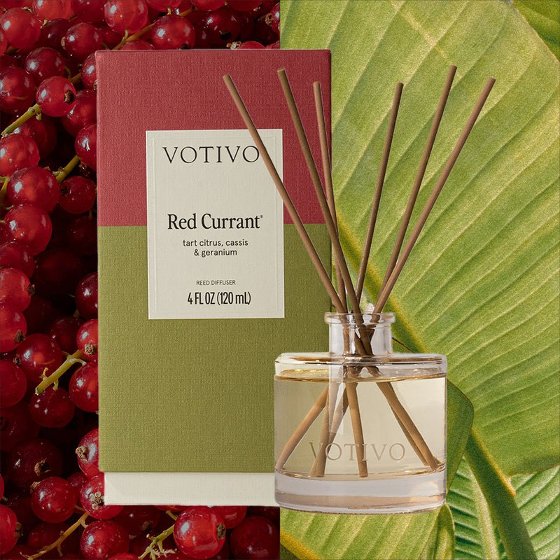 A Votivo - Red Currant Reed Diffuser, in its packaging, is displayed with red currants and green leaves. The clear bottle holds light liquid and brown reeds, capturing the scent of tart citrus and lush foliage.