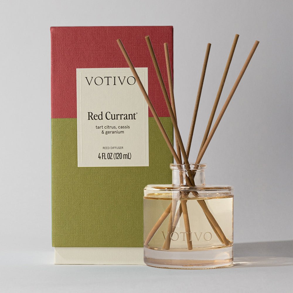 A Votivo - Red Currant Reed Diffuser with brown reeds stands in a clear glass bottle before its red and green box, set on a neutral background.