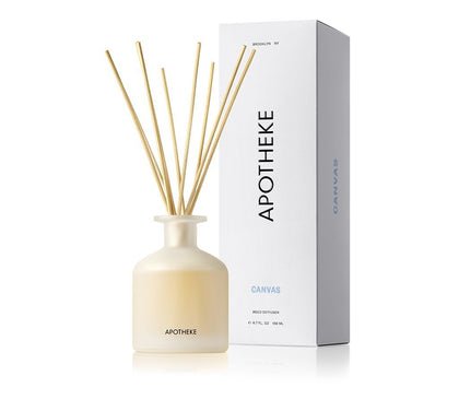 A frosted glass Apotheke - Canvas Reed Diffuser features slender wooden reeds that subtly release white musk notes. Its labeled Apotheke Canvas, and a matching rectangular box with the same branding stands beside it.