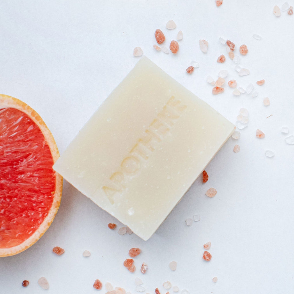 The Apotheke - Sea Salt Grapefruit Bar Soap rests on a light surface with pink sea salt grains scattered around, and a half grapefruit partially visible on the left, indicating its refreshing citrus fragrance.