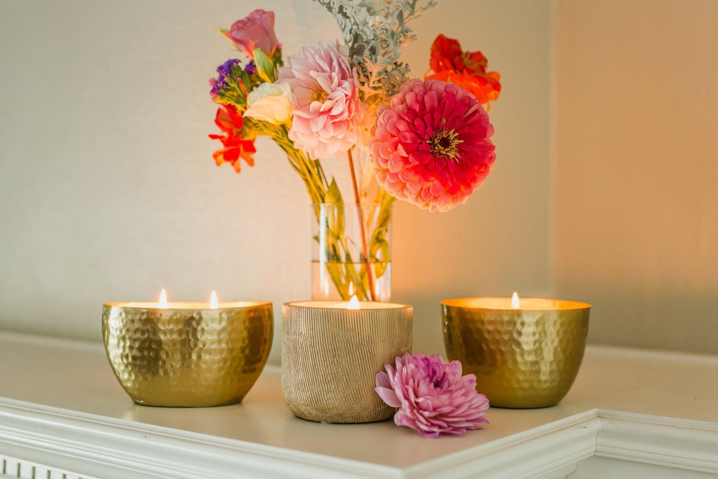 On a white surface, three lit Aqua De Soi - Amber and Teakwood candles fill the air with teakwood and golden amber. Behind them, a vase holds vibrant pink and orange flowers, while a pink flower lays nearby, adding to the cozy ambiance.