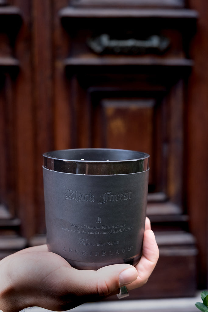 A hand holds the Archipelago - Black Forest Letter Press 13oz Candle with a metallic finish and engraved text, in front of a decorative wooden door. Hints of ebony woods add depth to this sophisticated Archipelago design.