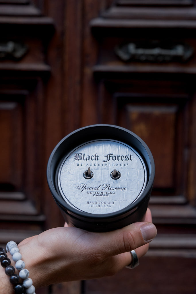A hand holds the Archipelago - Black Forest Letter Press 13oz Candle with two unlit wicks. The scent evokes ebony woods, while a carved brown wooden door in the background adds rustic charm.