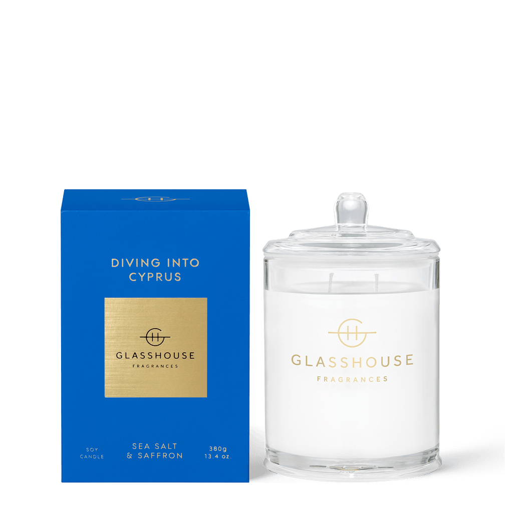 A blue box with gold lettering reads Diving Into Cyprus. Beside it is a 13.4oz Glasshouse Fragrances Triple Scented Soy Candle in a clear jar with lid, featuring SEA SALT & SAFFRON notes and the brands logo, all set against a crisp white background.