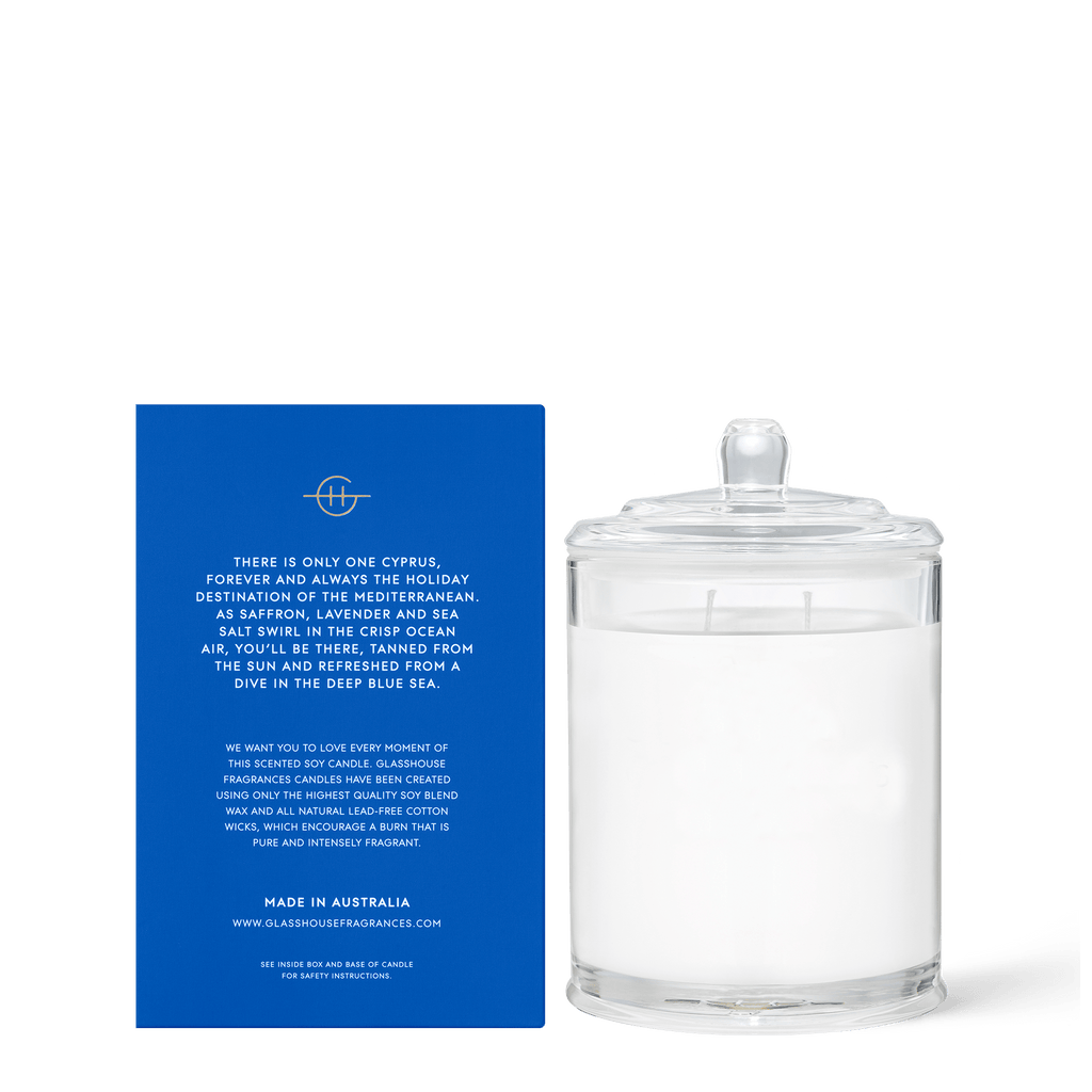 The Glasshouse Fragrances - Diving into Cyprus 13.4oz Candle, made from non-toxic soy blend wax, sits beside a blue box with white text detailing its inspiration and origin, including Cyprus and the Mediterranean. This unlit SEA SALT & SAFFRON candle is triple scented and appears clear in color.