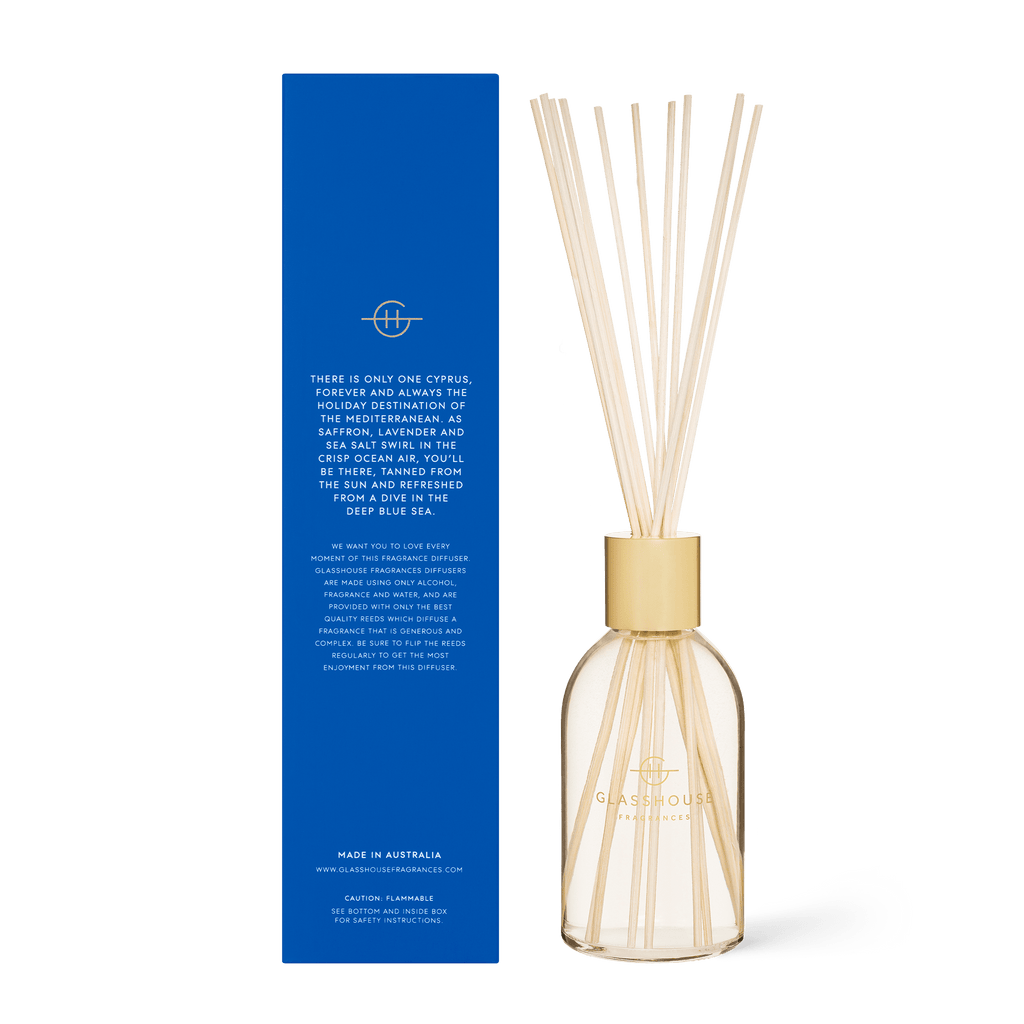 The Glasshouse Fragrances - Diving into Cyprus Diffuser features a glass bottle with a wooden cap and reeds. Its elegantly displayed beside a tall blue box with white and gold text, describing the SEA SALT & SAFFRON scent inspired by the Mediterranean Sea—offering a refreshing, non-toxic soy blend.