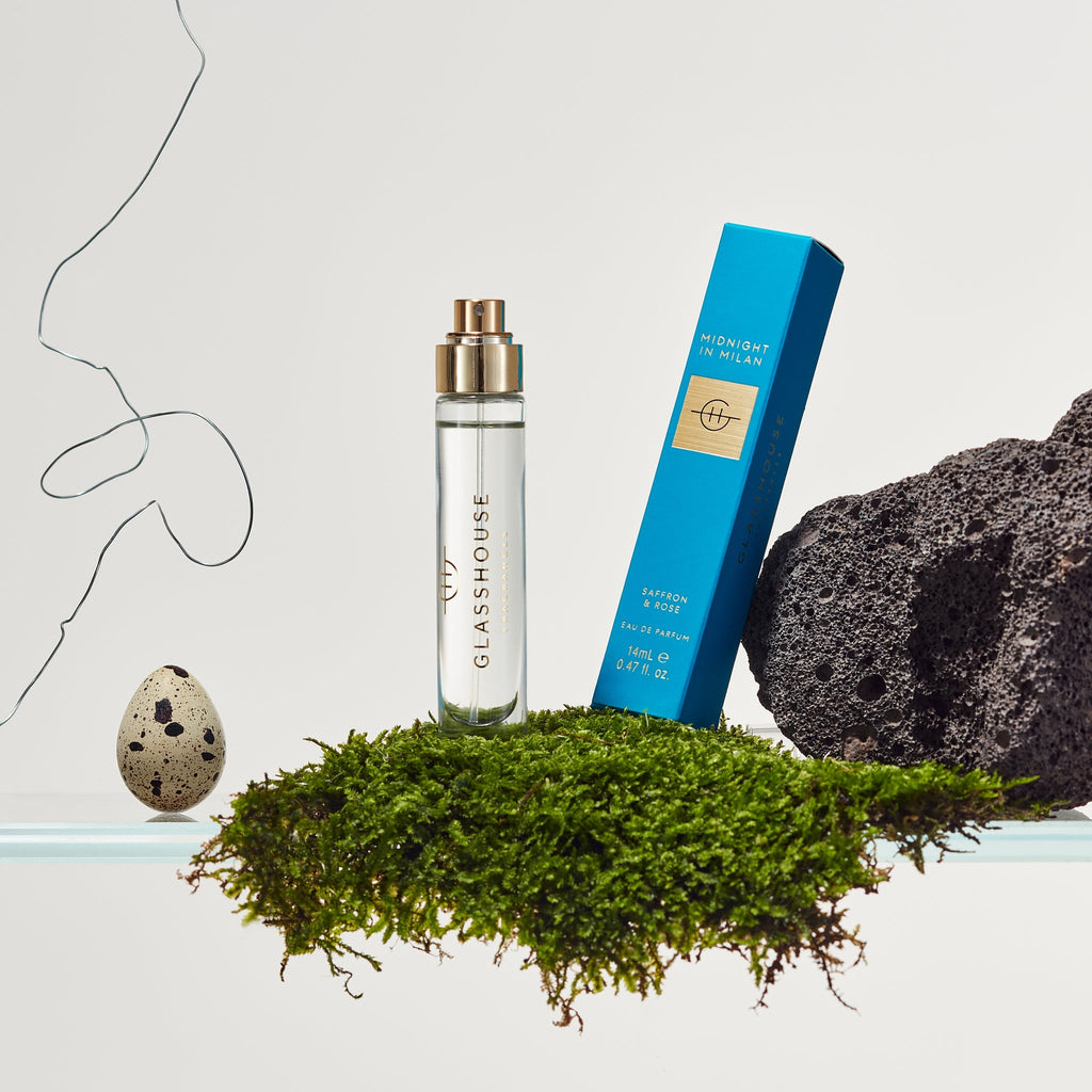 The Glasshouse Fragrances Eau de Parfum 14ml – Midnight In Milan bottle stands upright beside its blue box on green moss, with a speckled egg and dark volcanic rock, set against a light backdrop with subtle line art.