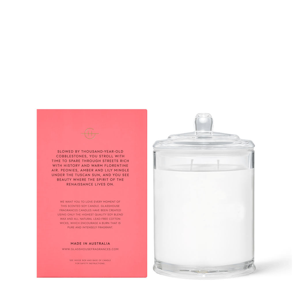 The Glasshouse Fragrances - Forever Florence 13.4oz Candle, a two-wick glass jar candle, sits beside a coral box with elegant text. This triple-scented Australian soy candle exudes luxurious, hand-crafted quality and fills the air with a hint of peonies.