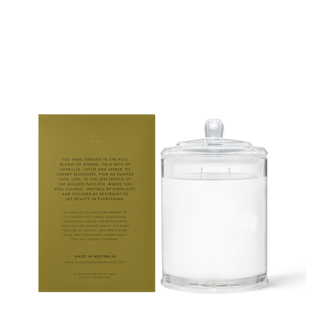 The Glasshouse Fragrances - Kyoto in Bloom 13.4oz Candle, in clear glass with a lid, sits next to its olive-green box adorned with gold Camellia & Lotus fragrance details, both displayed on a plain white background.