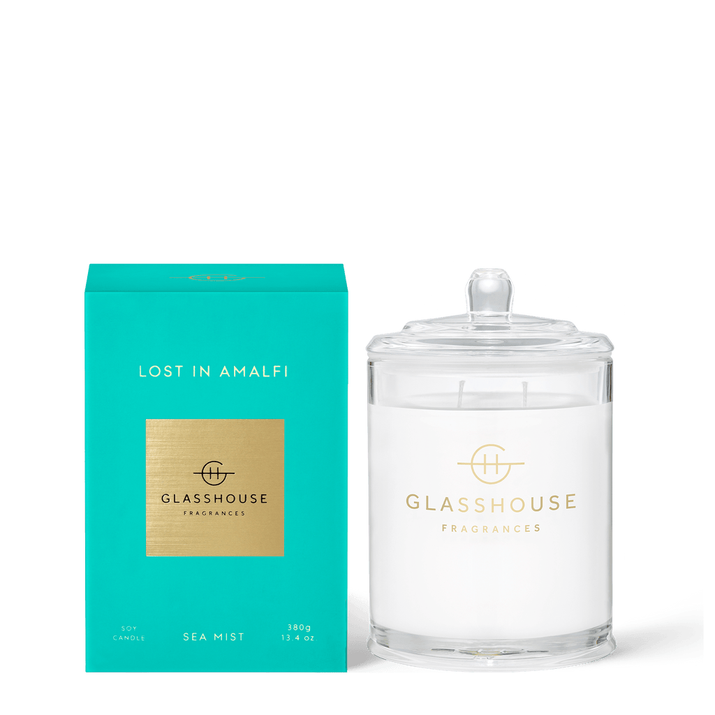 The Glasshouse Fragrances - Lost in Amalfi 13.4oz Candle rests elegantly by its teal box, featuring a SEA MIST soy composition with non-toxic wax. It is encased in clear glass adorned with a minimalist gold logo, offering a triple-scented ambiance.