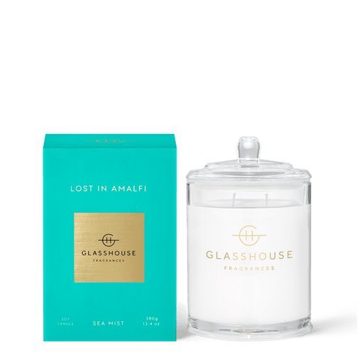 The Glasshouse Fragrances - Lost in Amalfi 13.4oz Candle rests elegantly by its teal box, featuring a SEA MIST soy composition with non-toxic wax. It is encased in clear glass adorned with a minimalist gold logo, offering a triple-scented ambiance.