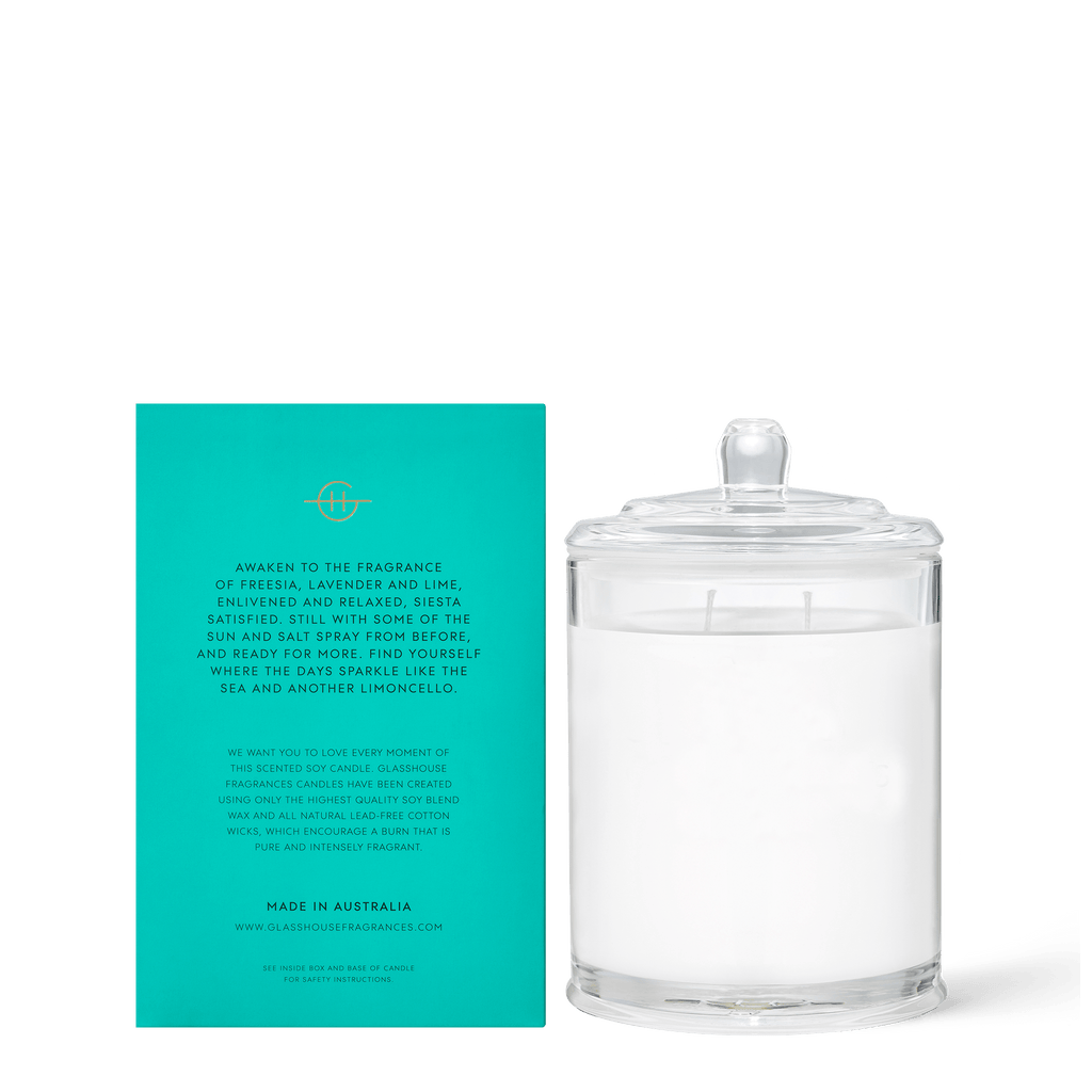 A Glasshouse Fragrances - Lost in Amalfi 13.4oz candle jar with a lid sits next to a teal card detailing its triple-scented, non-toxic white wax and two wicks.