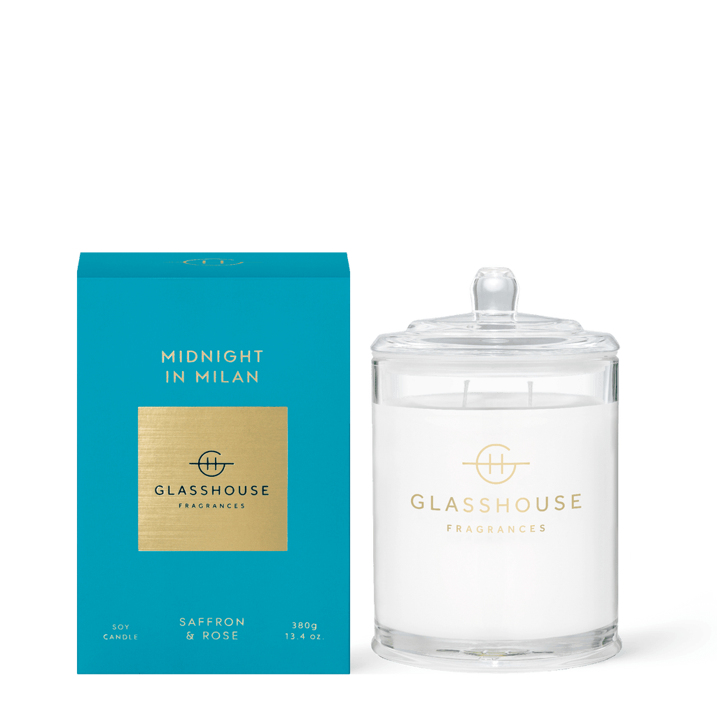 A Glasshouse Fragrances - Midnight In Milan 13.4oz Candle with a clear glass jar and lid is shown beside its turquoise and gold box, featuring a saffron and rose scent.