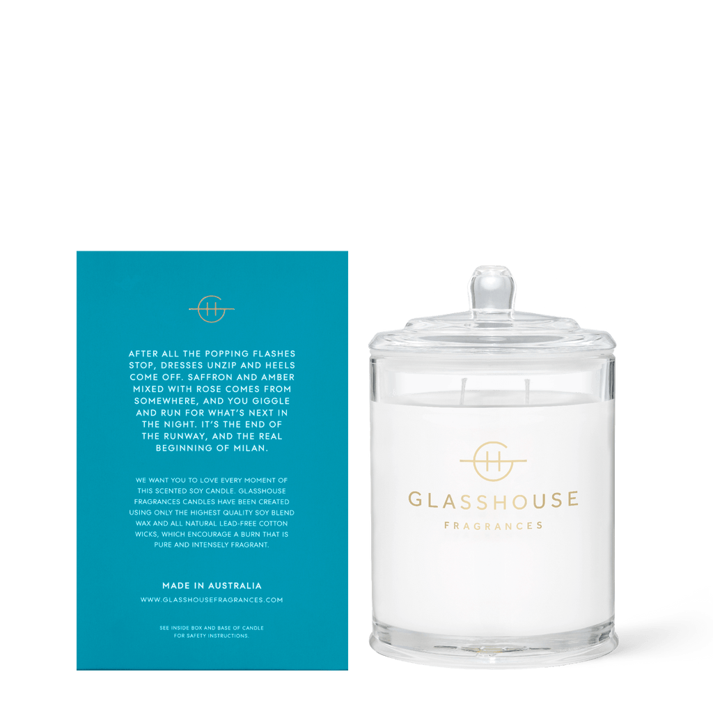 A Glasshouse Fragrances Midnight In Milan 13.4oz soy candle in a clear glass jar with lid sits beside a blue card with gold and white text detailing the brand’s inspiration, origin, and website.