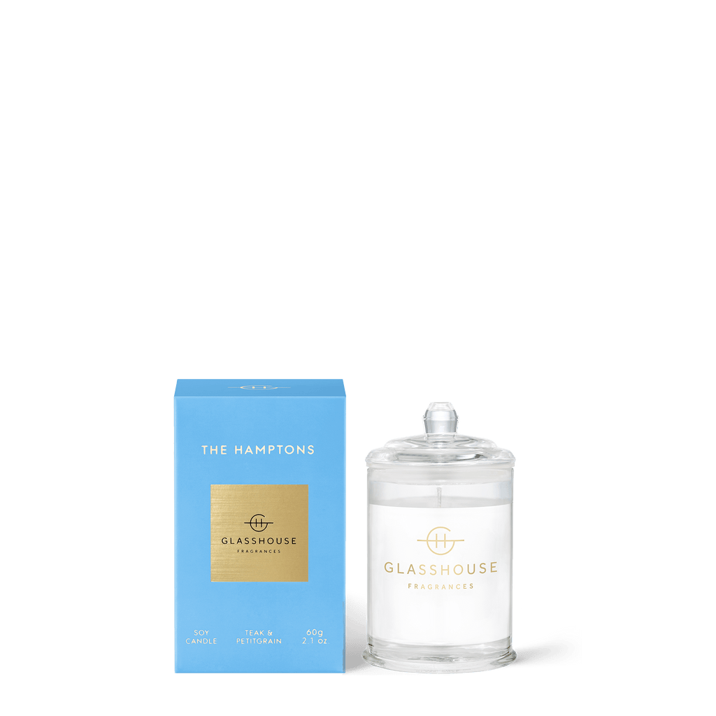 The Hamptons Mini 2.1oz Candle by Glasshouse Fragrances is crafted from natural, non-toxic wax, nestled in a clear glass jar with a lid, and comes in a blue box labeled Teak & Petitgrain, offering an indulgent aromatic experience.