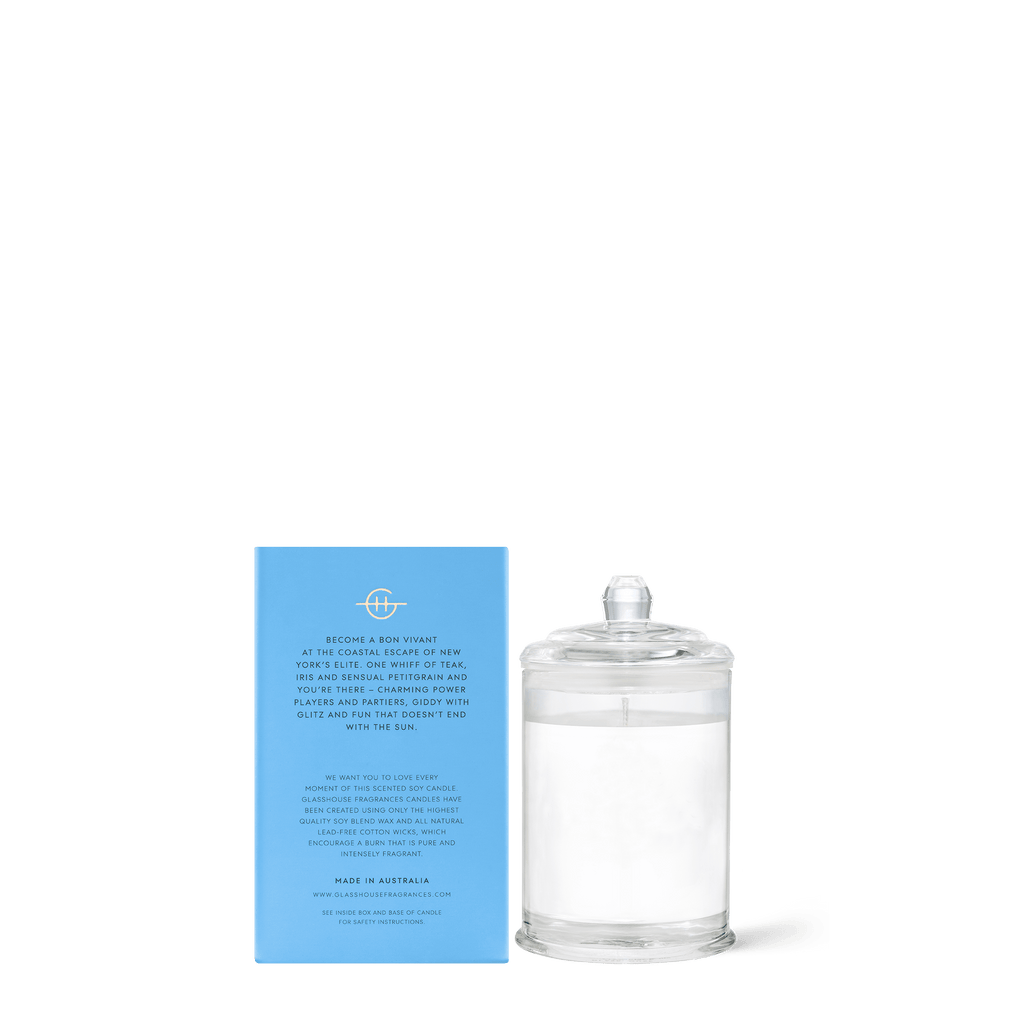 A clear glass candle with a white wick sits beside a light blue rectangular package. This Glasshouse Fragrances - The Hamptons Mini 2.1oz Candle offers Teak & Petitgrain scents, with Natural Lead-Free Cotton Wicks highlighted on the package along with other details.