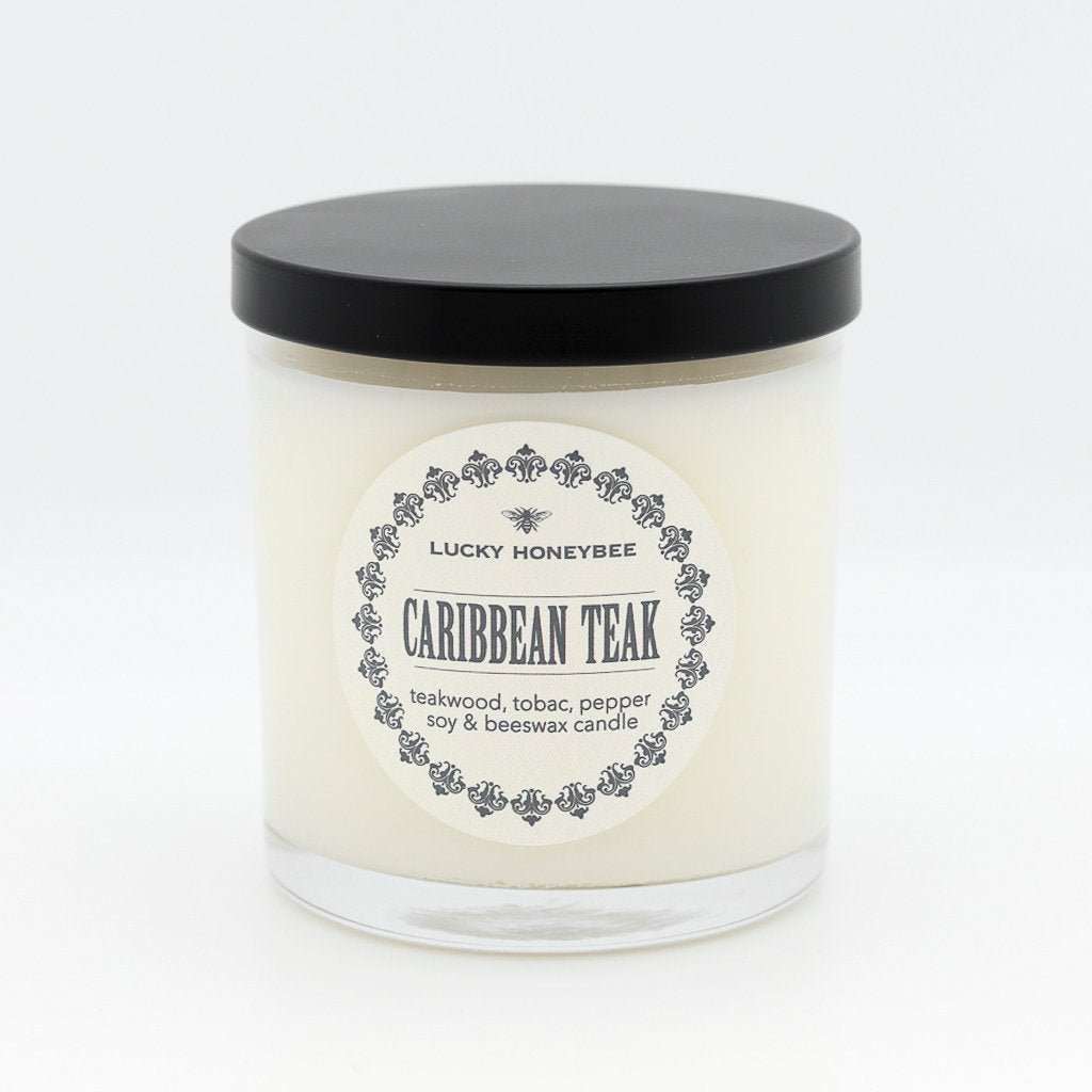 The Lucky Honeybee - Caribbean Teak Candle features soy and beeswax in a clear glass jar with a black lid, blending teakwood, tobac, pepper, and warm sandalwood notes for an elegant touch.
