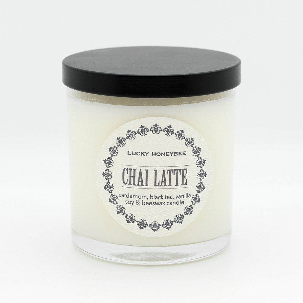 The Lucky Honeybee - Chai Latte Candle features a white jar with a black lid and a label displaying Lucky Honeybee and Chai Latte. It lists cardamom pods, black tea, vanilla, soy, and beeswax as ingredients. The design is elegantly set against a plain white background.