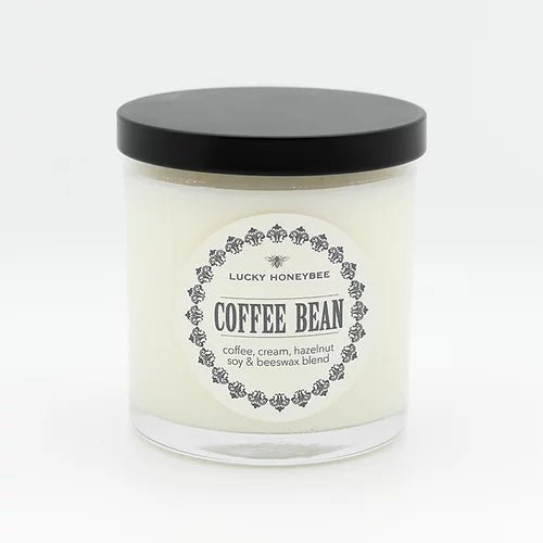 The Lucky Honeybee - Coffee Bean Candle features a white candle in a clear glass jar with a black lid. Its label, adorned with decorative black details, highlights the rich aroma of coffee, cream, and hazelnut. Made from a soy & beeswax blend and infused with essential oils.