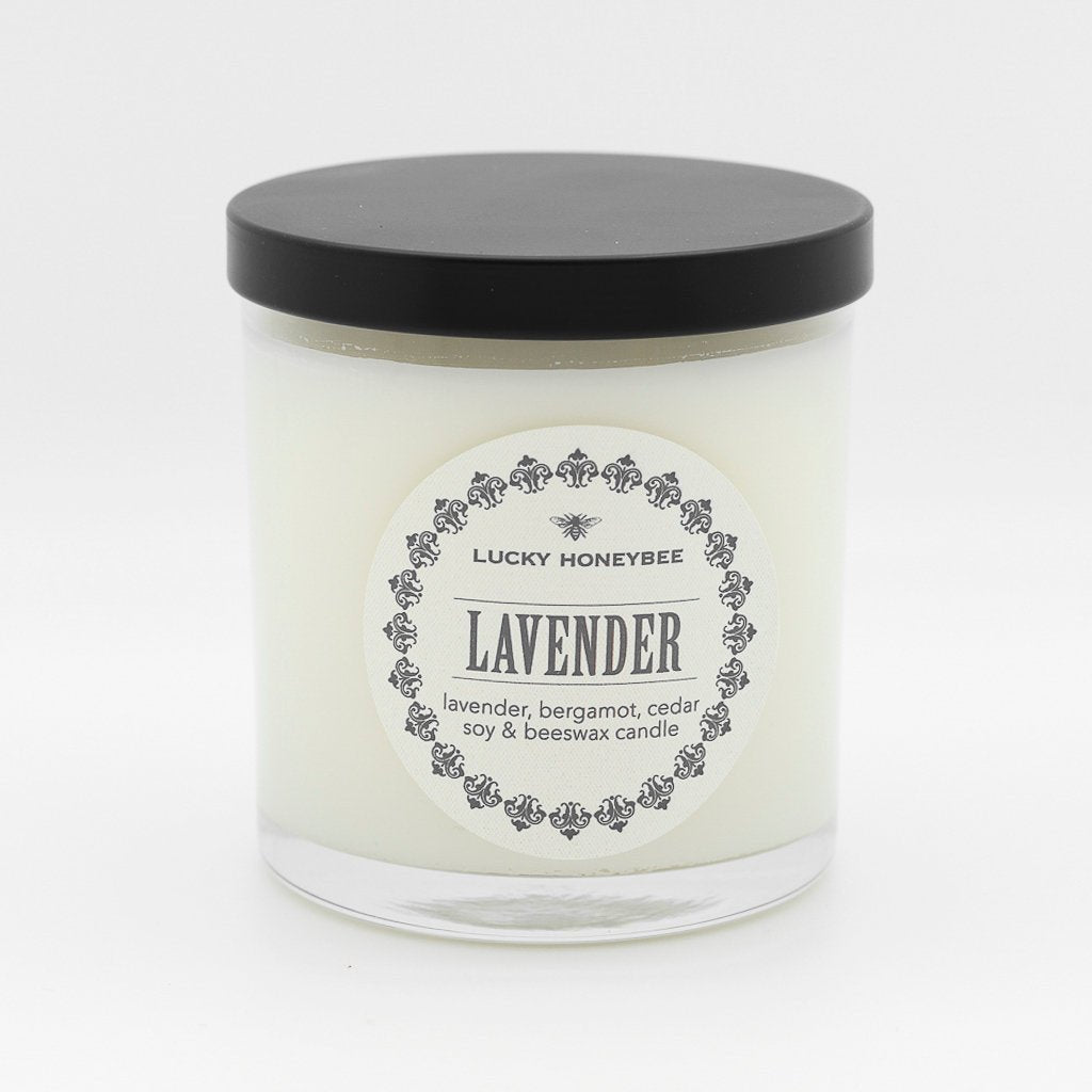 The Lucky Honeybee - Lavender Candle features a white soy and beeswax blend in a clear glass jar with a black lid. Its label, Lucky Honeybee Lavender, highlights soothing lavender, bergamot, and cedar scents, complemented by bee motif designs.