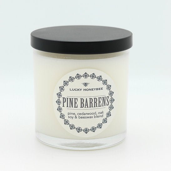 The Lucky Honeybee Pine Barrens Candle, by Lucky Honeybee, is a white soy and beeswax candle in a transparent glass jar with a black lid. It features spruce, pine, cypress, cedarwood, and oak notes on its label, perfect for Jersey expats seeking a woodland escape.