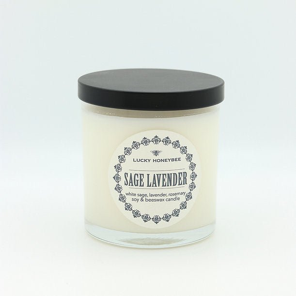 The Lucky Honeybee Sage Lavender Candle is a white candle in a glass jar with a black lid. Its label lists ingredients like white sage, lavender, rosemary, soy, and beeswax. Infused with sandalwood, it provides soothing scents for a serene space.