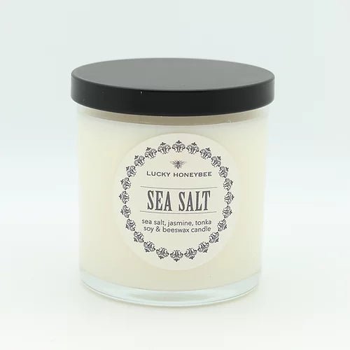 A white soy & beeswax candle by Lucky Honeybee in a clear glass jar with a black lid is labeled Lucky Honeybee Sea Salt. Featuring scents of sea salt, jasmine, and tonka, the labels text is framed by a decorative floral border.