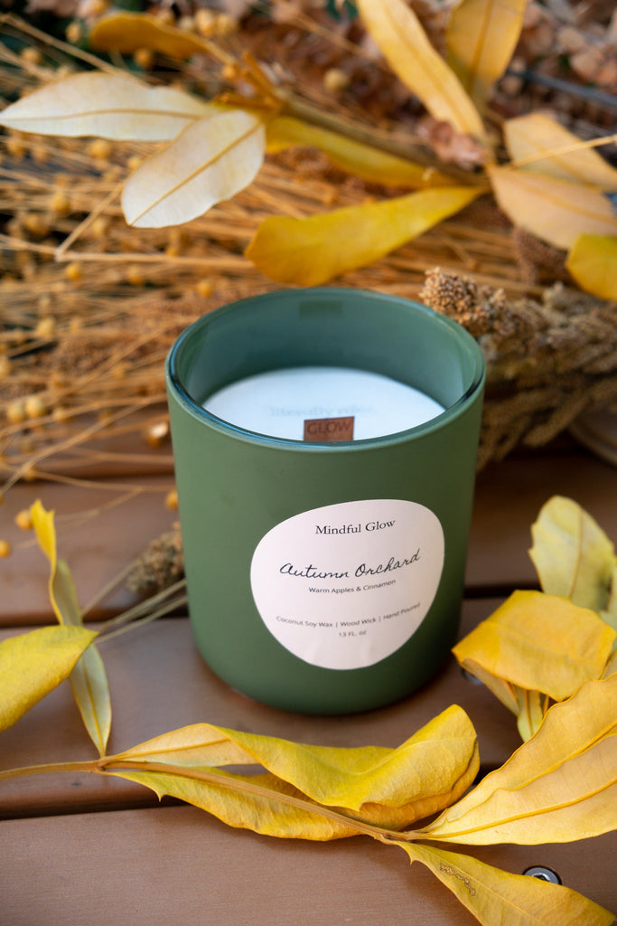 The Mindful Glow - Autumn Orchard Woodwick Candle 13oz rests unlit on a wooden surface, surrounded by yellow and brown leaves and dried foliage, evoking the fruity aroma of apple orchards.