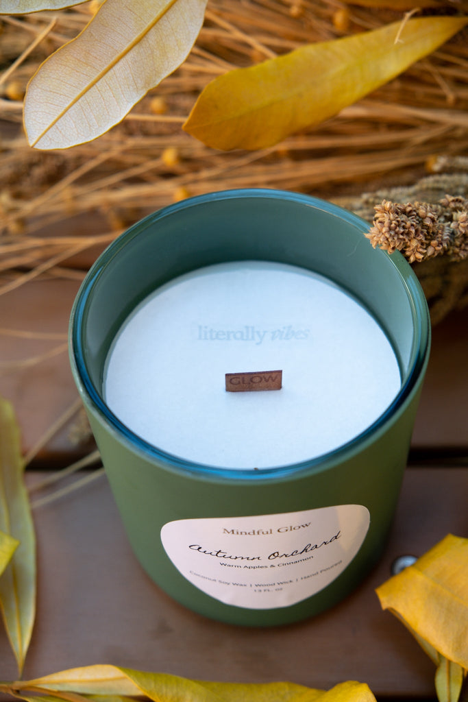 The Mindful Glow - Autumn Orchard Woodwick Candle 13oz sits among yellow leaves, its fruity aroma evoking apple orchards. A white lid and small brown GLOW tag accent the candle, while faint “literally vibes” text appears against wooden planks and dried leaves.