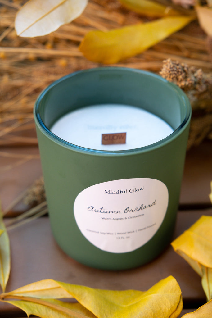 The Mindful Glow - Autumn Orchard Woodwick Candle 13oz evokes apple orchards with a fruity, warm apple and cinnamon scent, filling your space with autumn vibes as it sits among leaves and dried grasses.