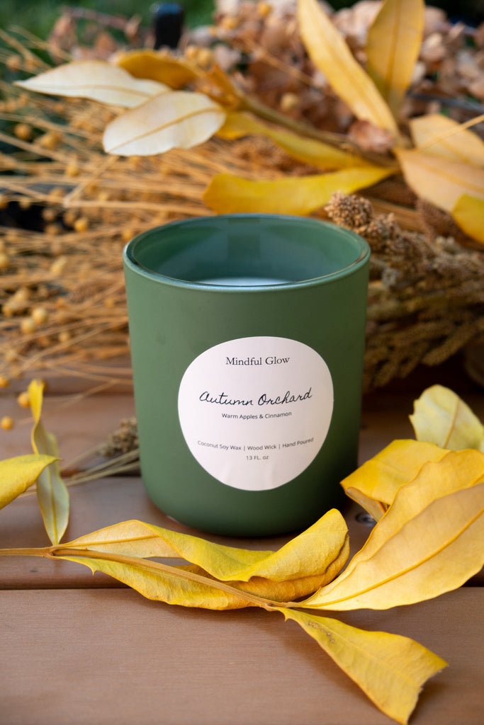 A Mindful Glow Autumn Orchard Woodwick Candle 13oz sits on wood among yellow leaves and dried flowers. Its label highlights a warm apple and cinnamon scent, capturing the cozy essence of fall apple orchards.