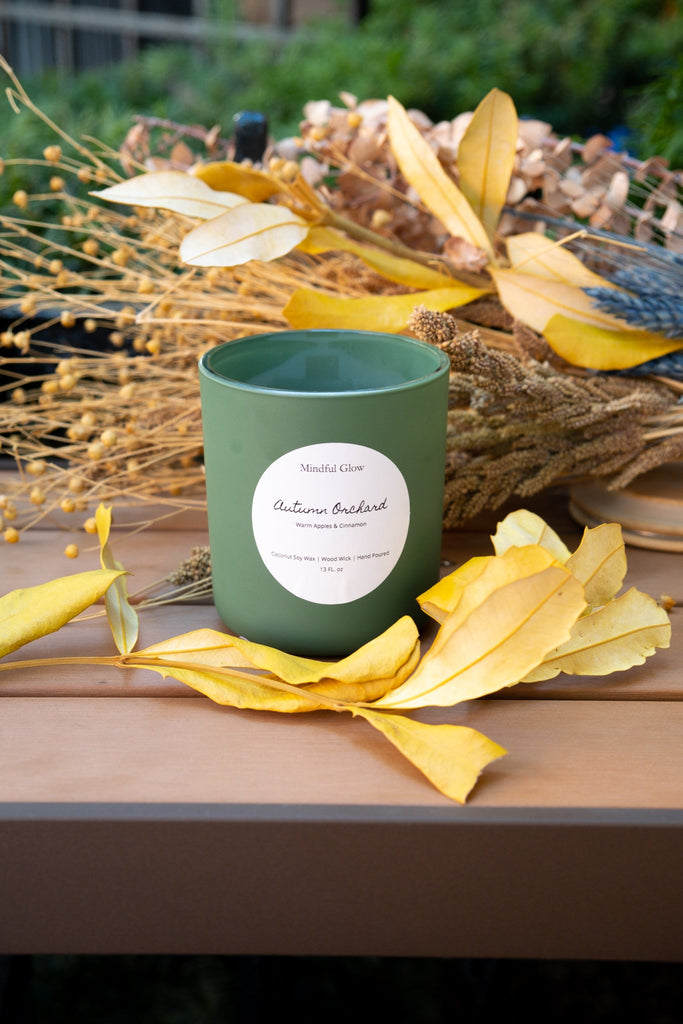 A Mindful Glow Autumn Orchard Woodwick Candle 13oz sits on a wooden table among yellow leaves and dried florals, filling the air with a fruity apple scent reminiscent of fresh orchards.