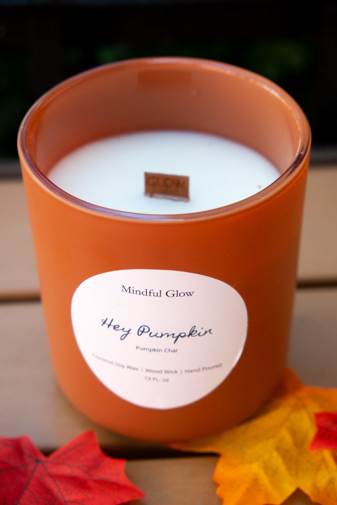 The Mindful Glow - Hey Pumpkin Woodwick Candle 13oz rests on a wooden surface with faux autumn leaves. Hand-poured coconut soy wax and a wood wick release a cozy pumpkin chai scent, bringing fall vibes from Mindful Glow.
