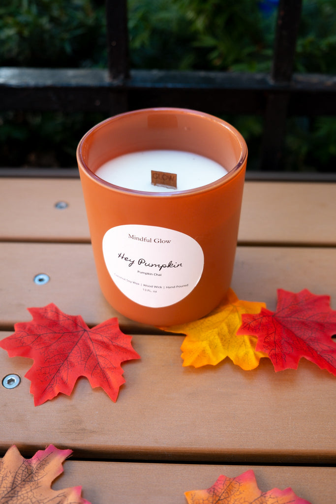 The Mindful Glow - Hey Pumpkin Woodwick Candle 13oz by Mindful Glow, made with coconut soy wax, sits on a wooden surface among artificial autumn leaves, with greenery and a black railing in the background.