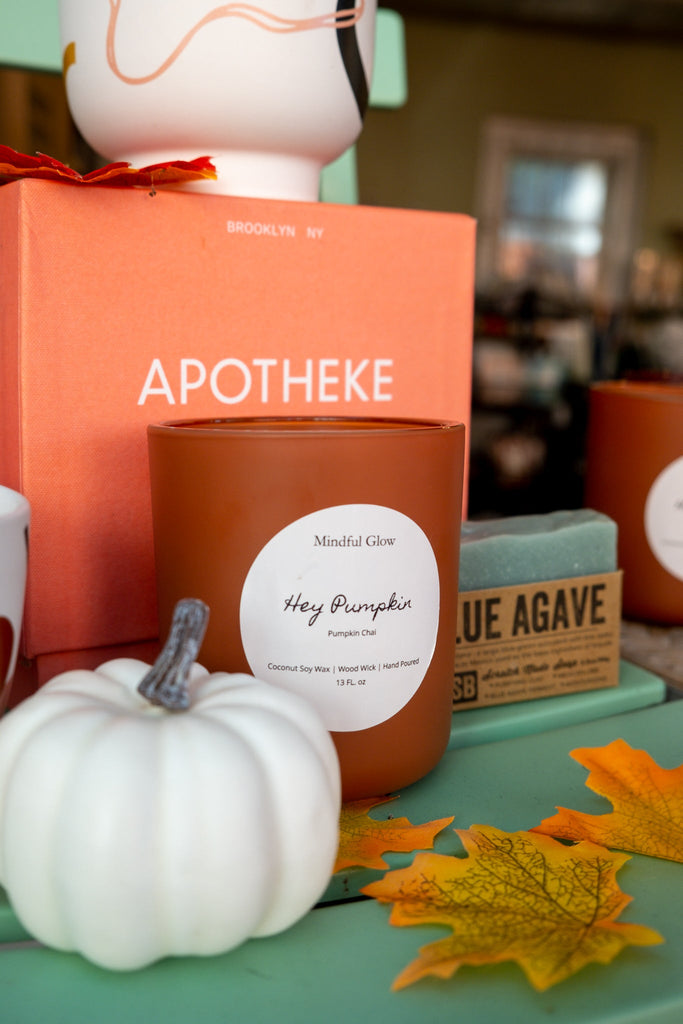 A 13oz Mindful Glow - Hey Pumpkin Woodwick Candle sits on a green surface next to a small white pumpkin, blue agave soap bar, orange box, and autumn leaves, creating a cozy fall display.