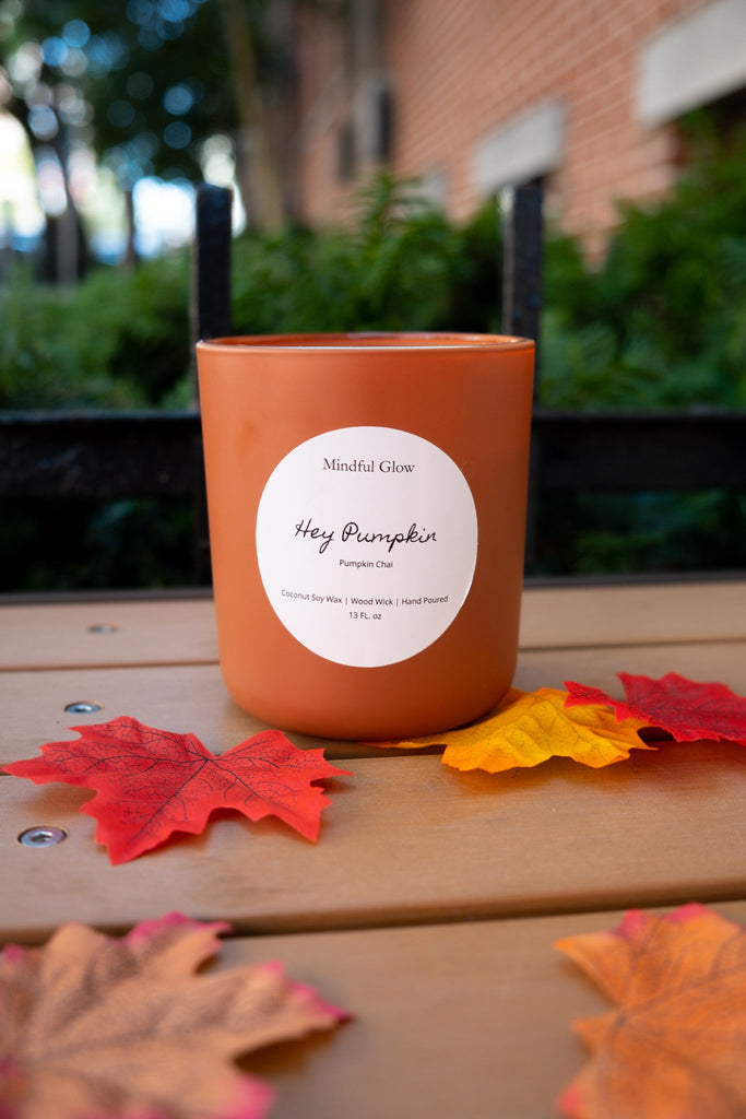 The Mindful Glow Hey Pumpkin Woodwick Candle 13oz sits on a wooden bench amid autumn leaves, with a green shrub and brick wall softly blurred in the background.