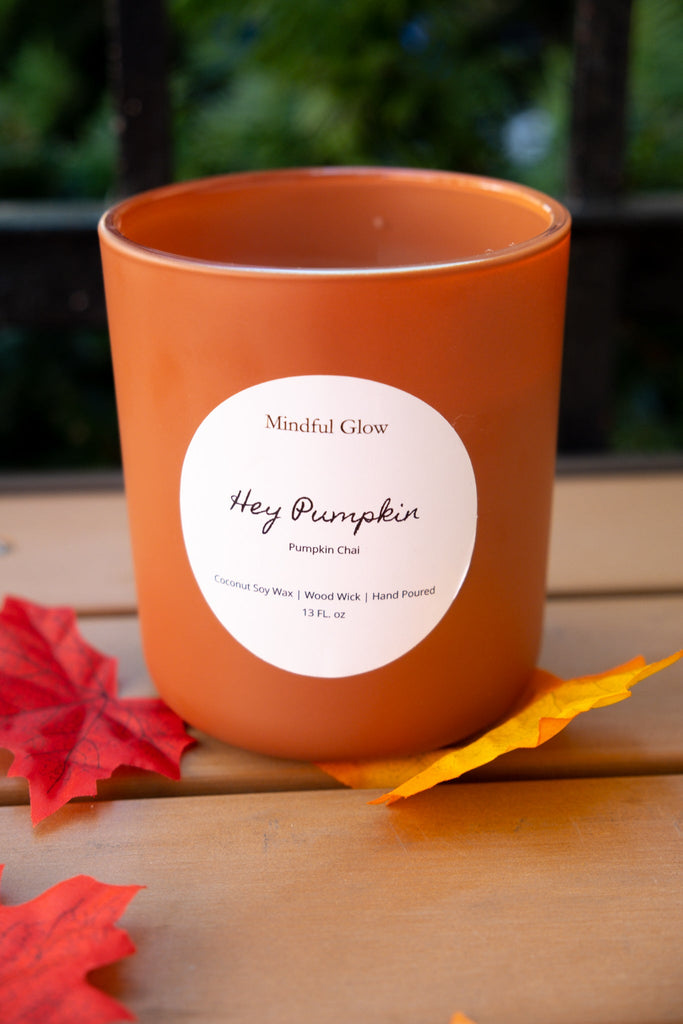 The Mindful Glow - Hey Pumpkin Woodwick Candle 13oz by Mindful Glow, made with coconut soy wax, sits on a wooden surface with red and yellow autumn leaves nearby; the background is softly blurred greenery.